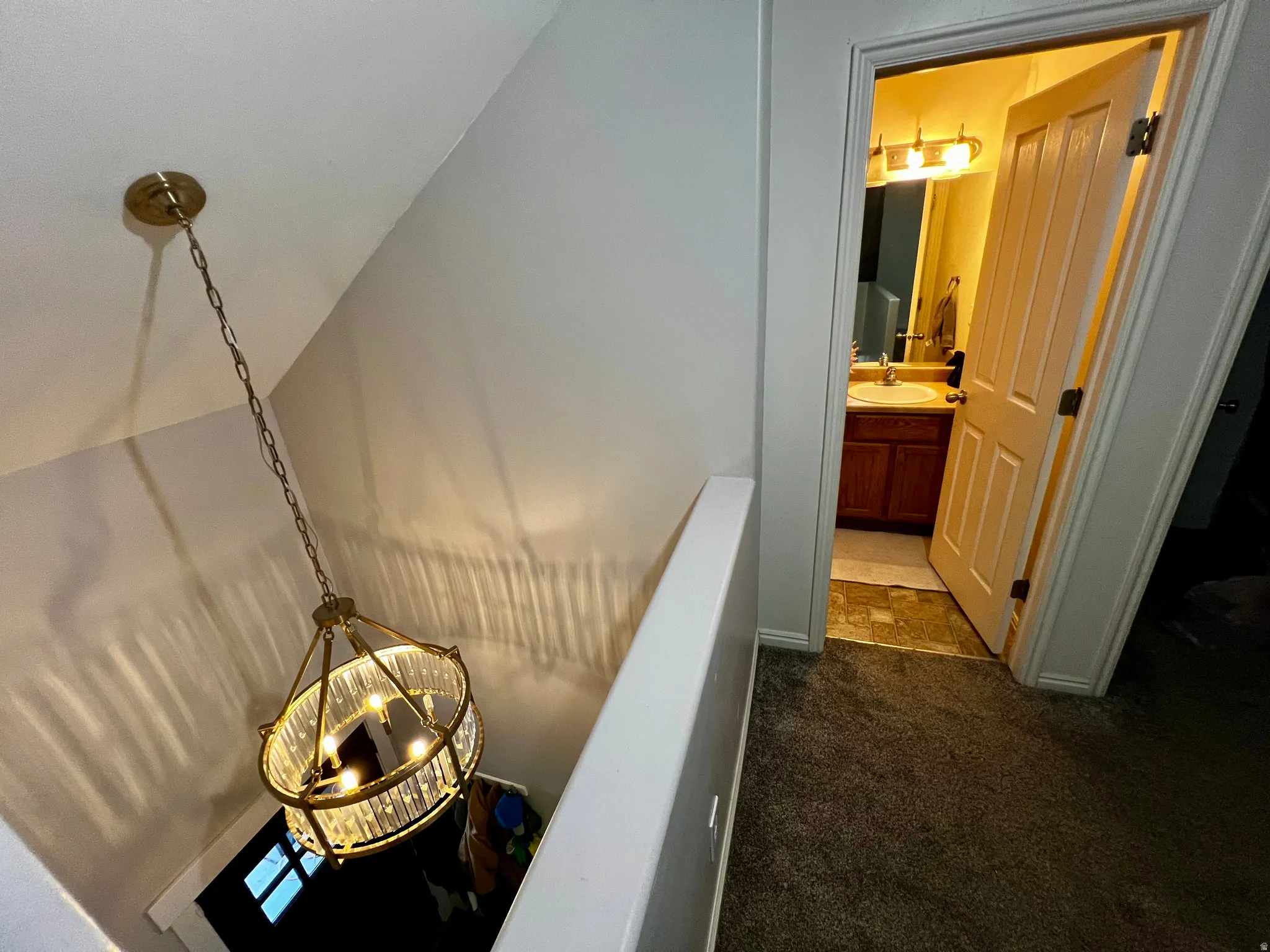 Crystal-adorned chandelier with a brass finish, carpeted landing, and a bathroom featuring a vanity with a wood-finish cabinet, countertop, and an overhead light fixture