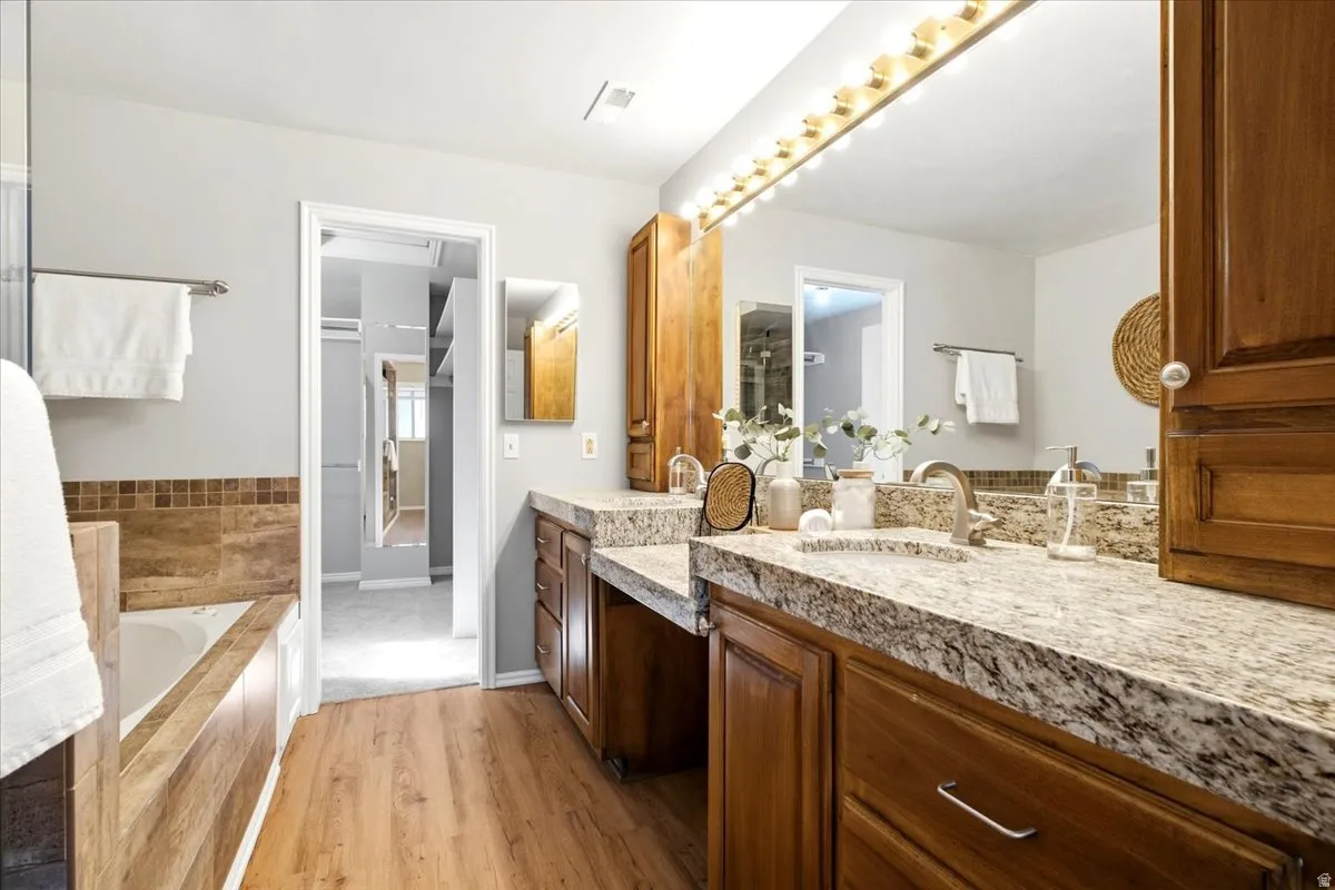 Dual vanity with undermount sinks and extensive countertop space, wood-finish flooring, built-in soaking tub, and natural wood cabinetry