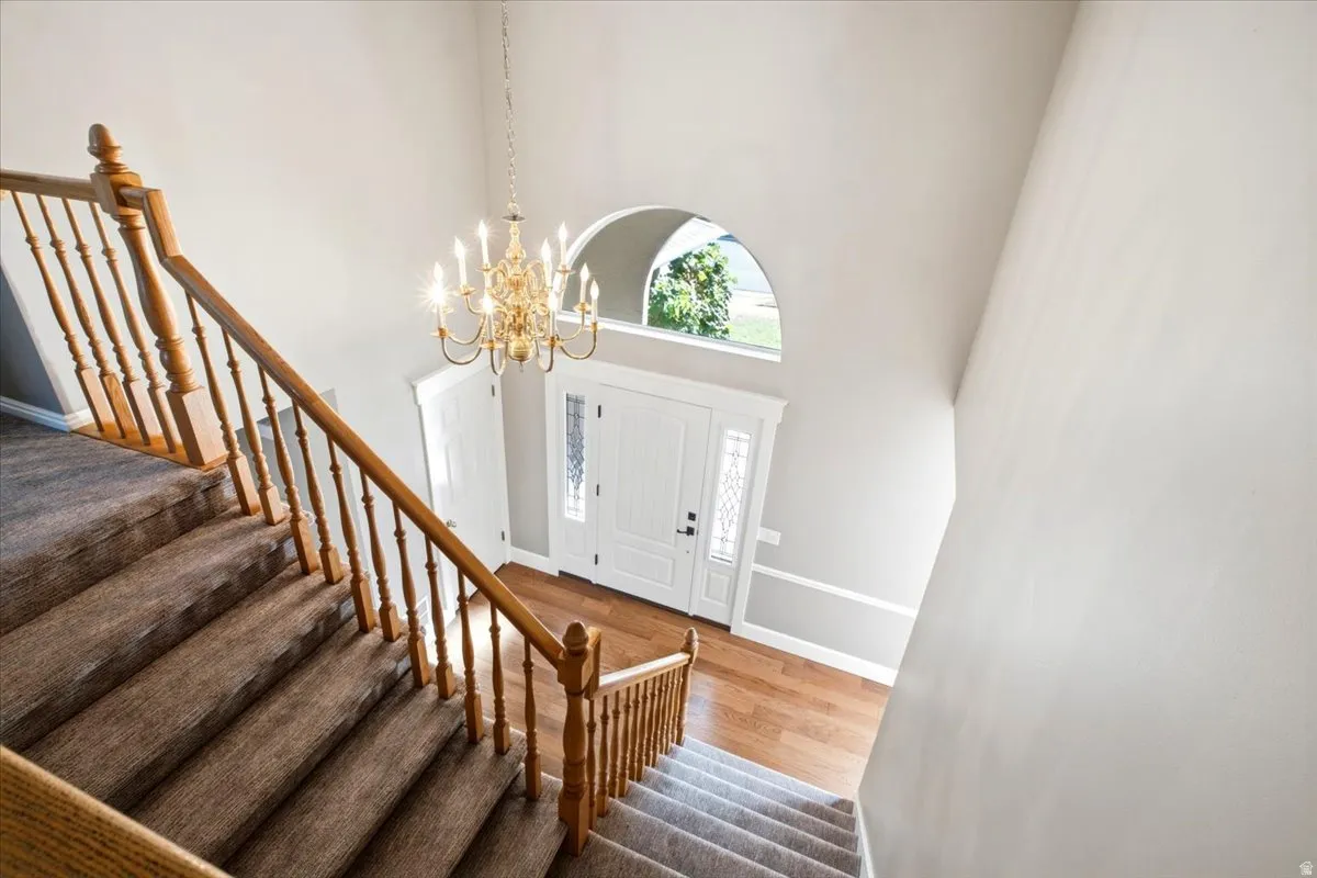 Carpeted staircase with wood banisters, prominent chandelier lighting, hardwood flooring at the entry, and an arched transom window above the main entry