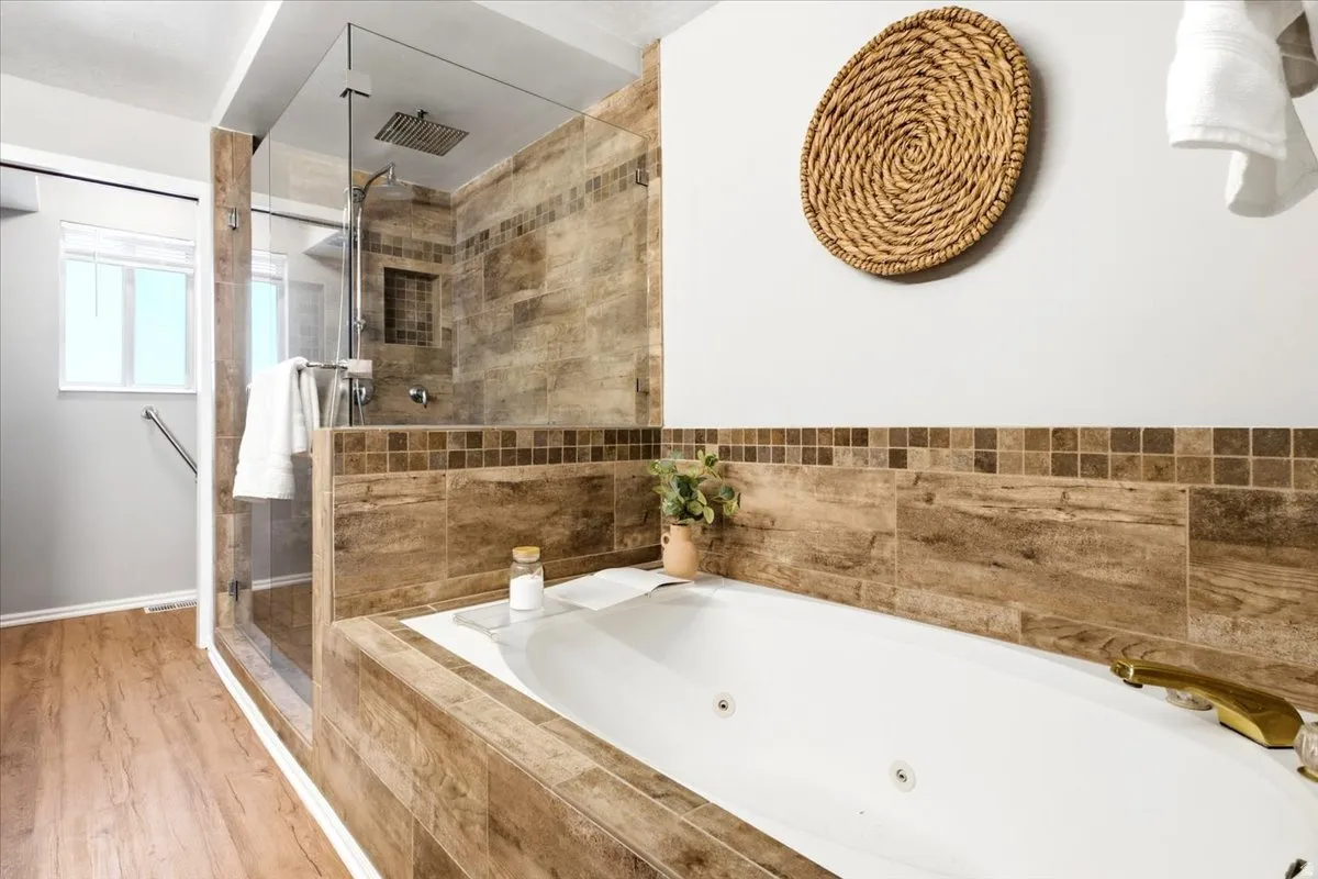 Jetted soaking tub with tile surround, walk-in shower with glass enclosure, wood-finish flooring, and a large window