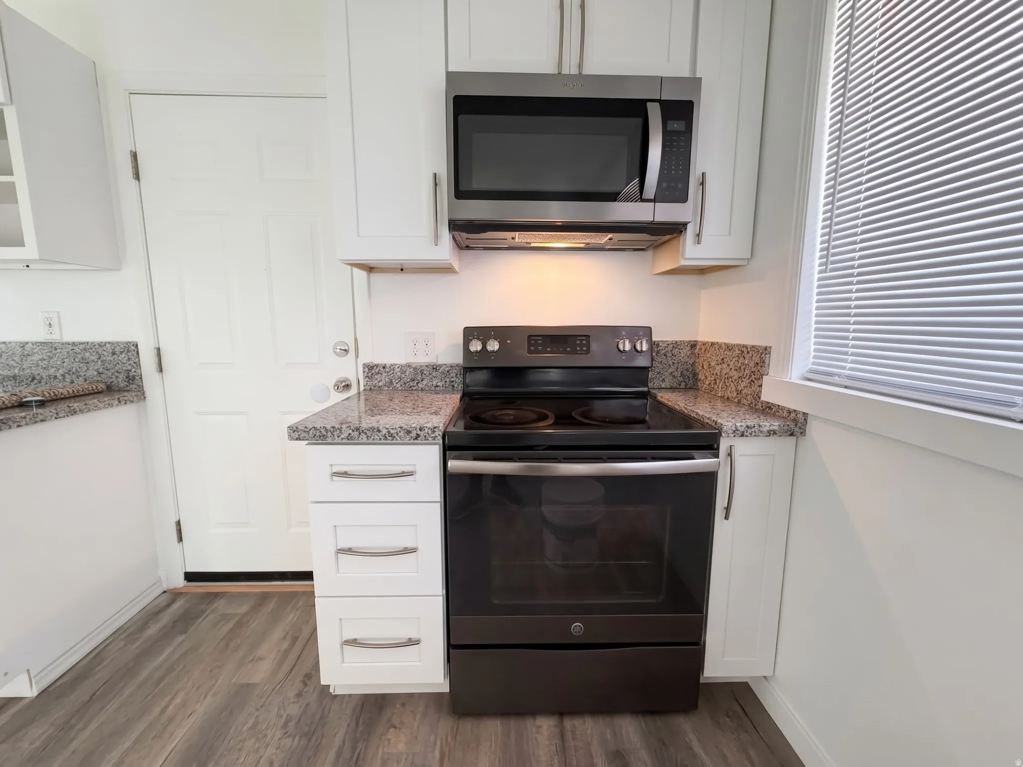 White shaker cabinetry, dark wood-finish flooring, black electric range, stainless steel over-the-range microwave, and granite countertops