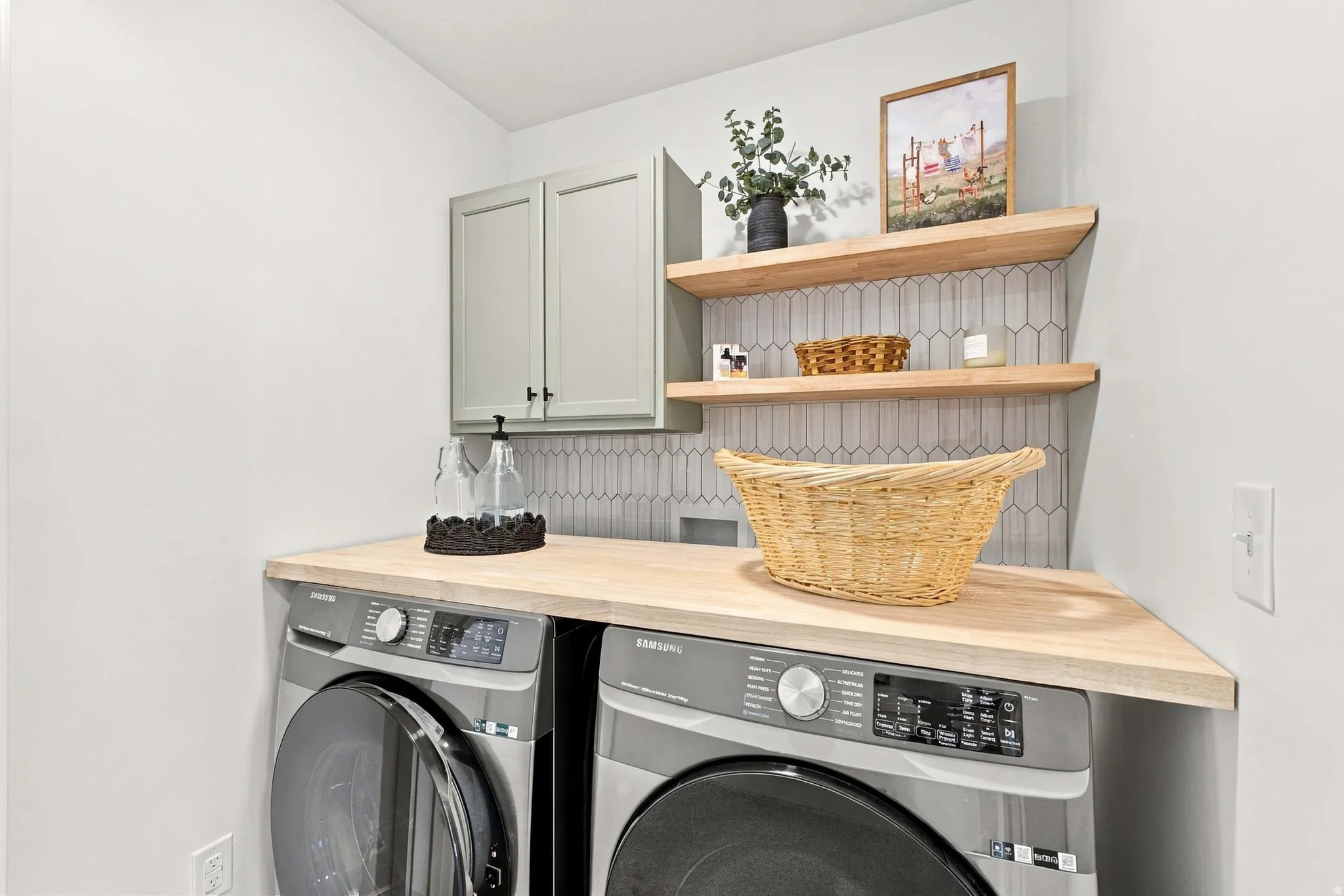 Light wood-finish countertop, floating wood-finish shelving, pale green shaker-style cabinetry, and a geometric tiled backsplash