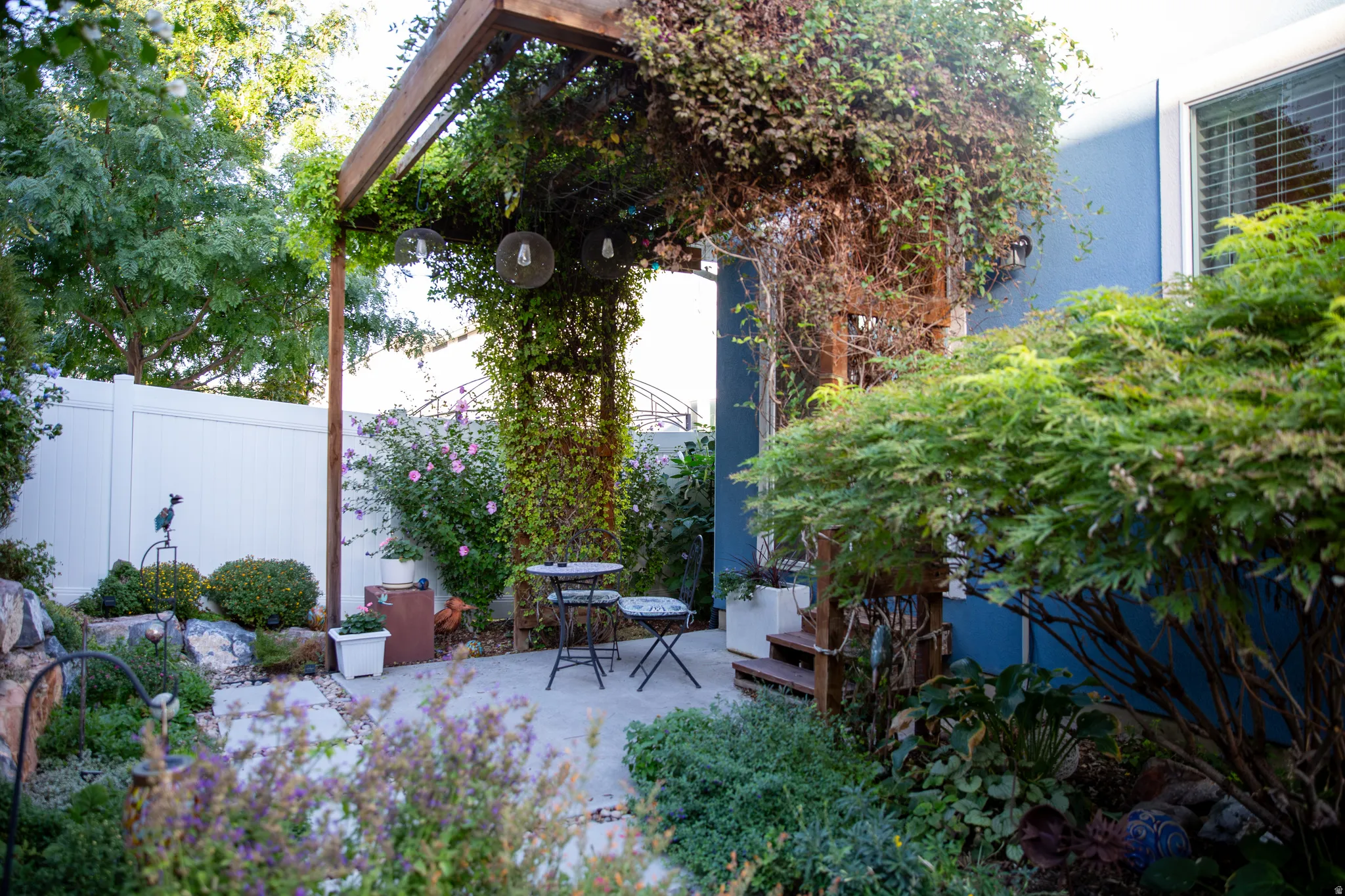 Paved patio with an overhead pergola, lush perennial plantings, mature trees, and a privacy fence