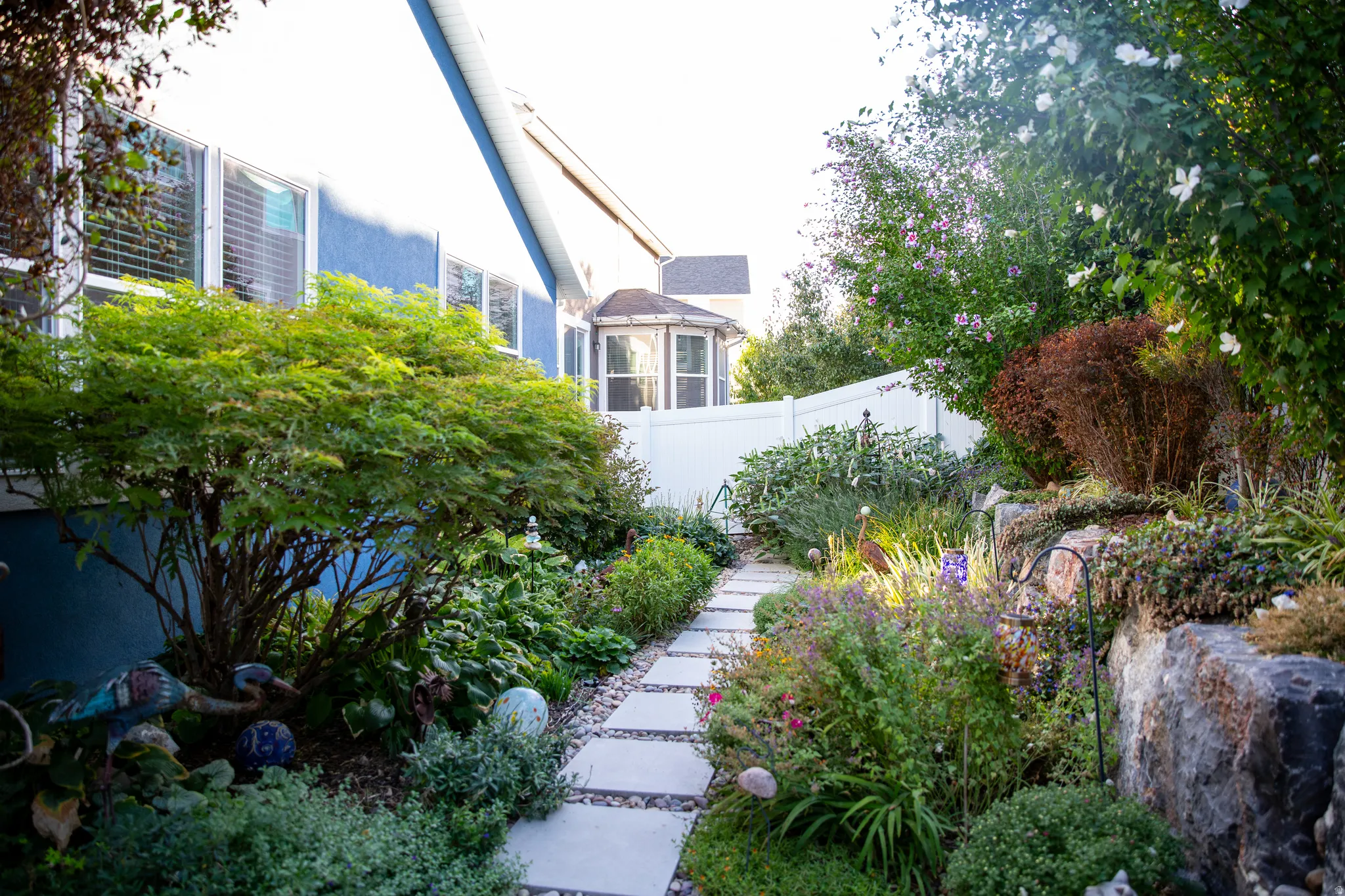 Stone paver pathway bordered by landscaped garden beds, featuring a rock retaining wall and a white vinyl privacy fence