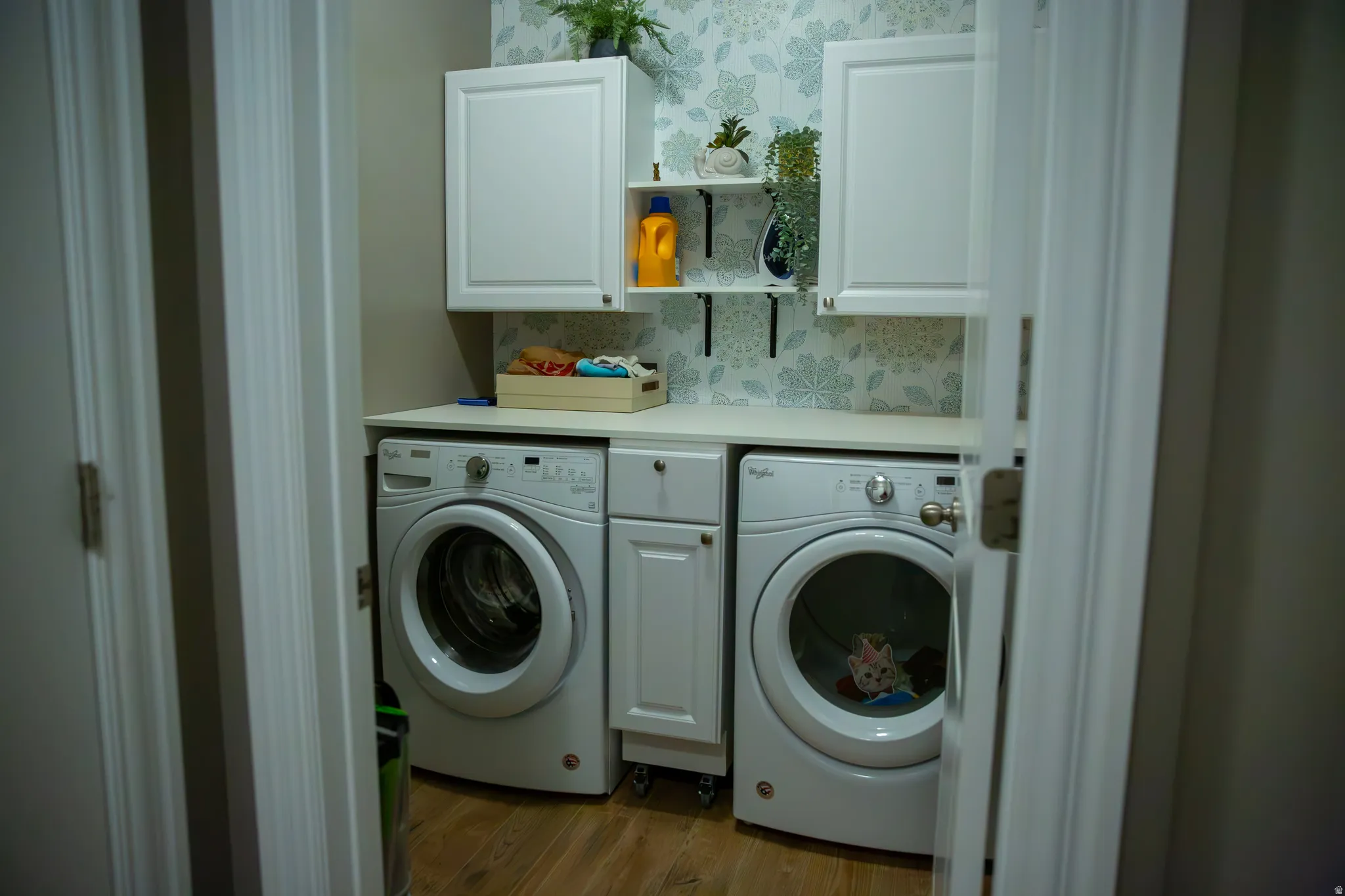 Laundry area with wood-finish flooring, white cabinetry, a light-toned countertop, and patterned wall covering