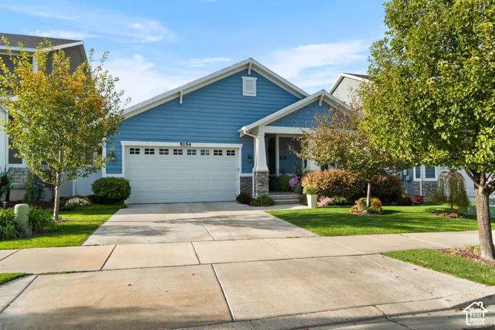 Blue siding exterior with white trim, a two-car garage with white door, a covered porch with stone accents, a concrete driveway, and a manicured lawn with mature trees