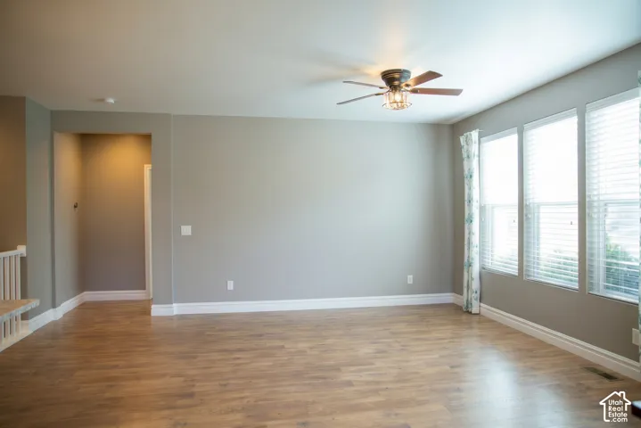 Wood-finish flooring throughout, light gray wall color, three large windows with blinds, ceiling fan with integrated lighting, and white baseboards