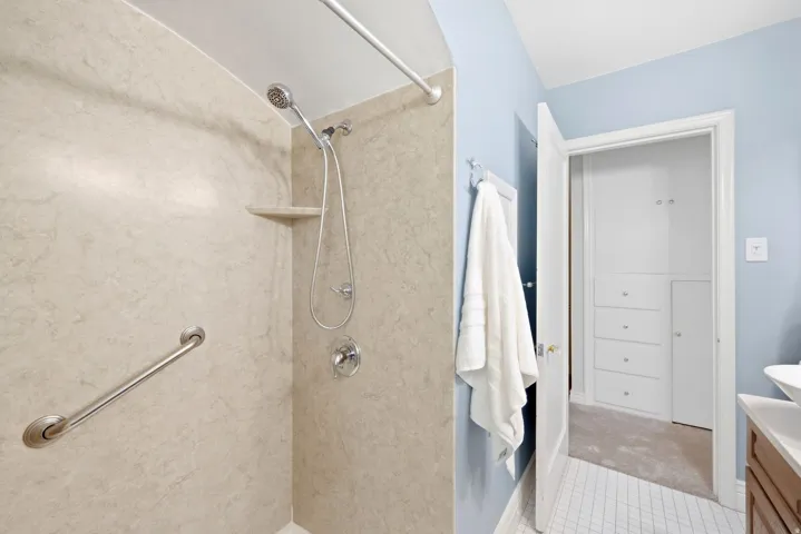 Shower stall featuring neutral-toned wall panels, a showerhead with an attached handheld sprayer, and a built-in grab bar