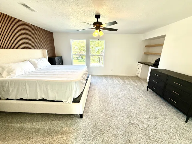 Plush carpeting, wood-paneled accent wall, dual windows, built-in desk with shelving, and overhead lighting with ceiling fan