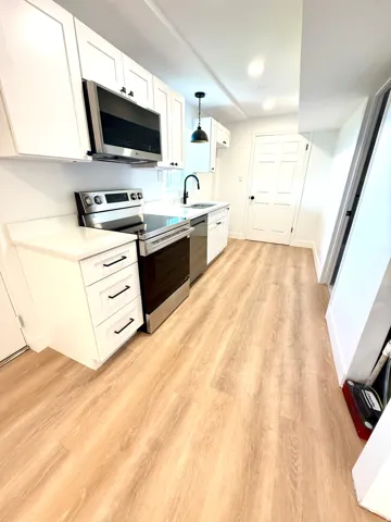 Galley kitchen featuring white cabinetry, stainless steel appliances, light-toned wood-finish flooring, and a contemporary pendant light fixture