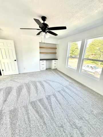 Recessed built-in desk with white cabinetry and dark countertop, wood shelving, overhead ceiling fan with light fixture, light gray carpeting, and two windows