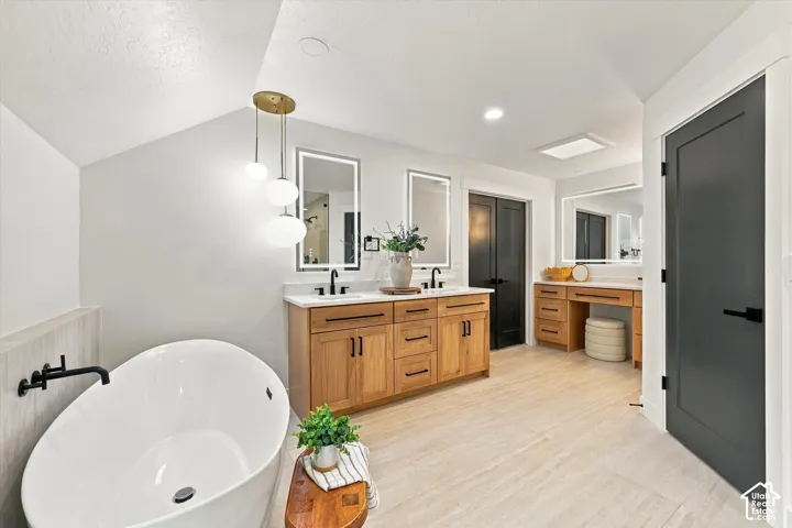 Freestanding soaking tub with a wall-mounted matte black faucet, dual wood-finish vanities with undermount sinks, and integrated mirror lighting