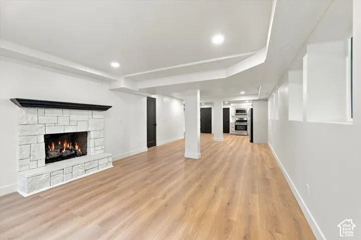 Stone-faced fireplace with a dark wood mantel, wood-finish flooring, recessed lighting, and a partial kitchen with stainless steel appliances