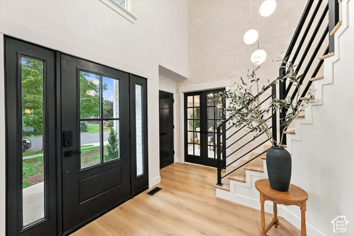 Double entry doors with glass panels and sidelights, light wood-finish flooring, two-story foyer with textured accent wall, open staircase with black metal railings, and double french doors