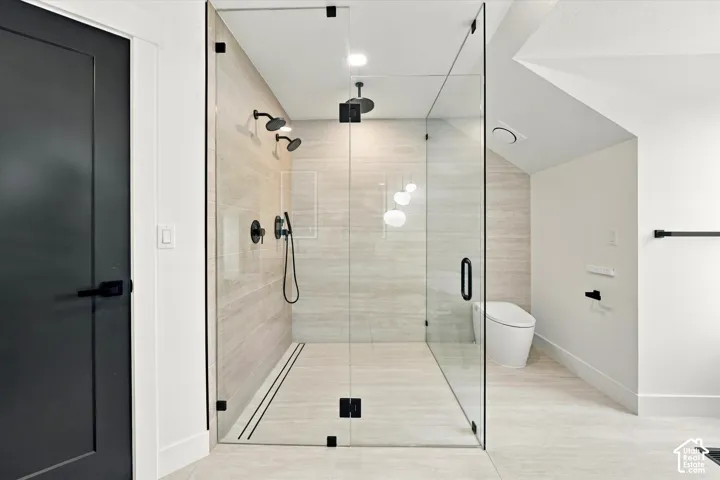 Glass-enclosed shower with multiple shower heads and matte black fixtures, light-toned tiled walls, light-toned floor tiling, and a black six-panel door