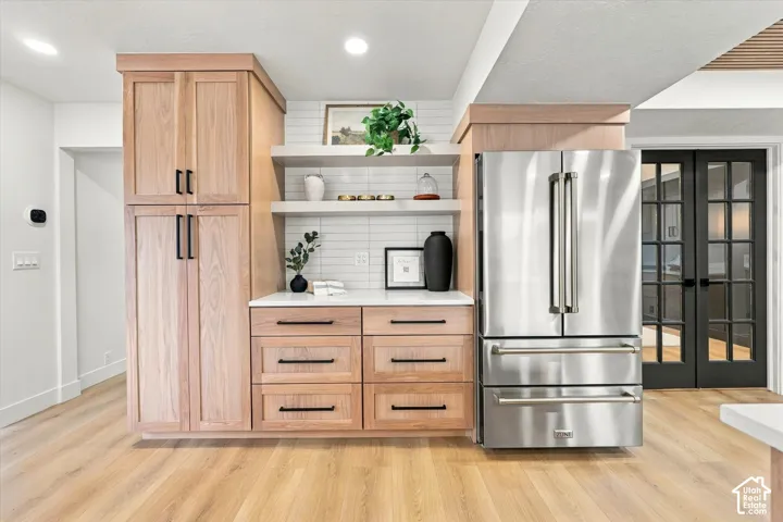 Wood-finish flooring with light wood cabinetry, stainless steel refrigerator, white subway tile backsplash, and dark framed French doors