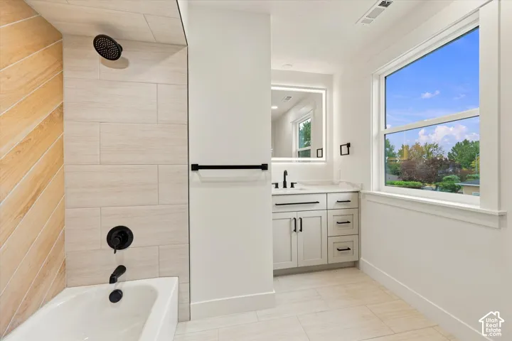 Bathtub with matte black fixtures, chevron-patterned shower wall, white vanity with matte black hardware, framed mirror with integrated lighting, and a large window