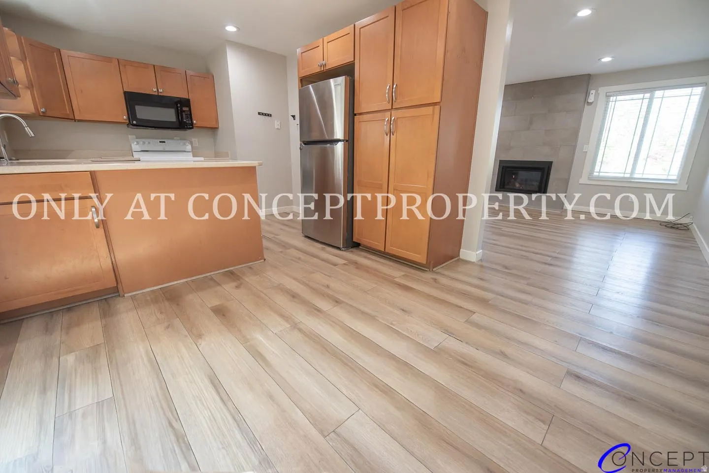Wood-finish flooring throughout, kitchen with wood cabinetry and stainless steel refrigerator, white countertops, built-in microwave, and a fireplace with a light-colored tile surround