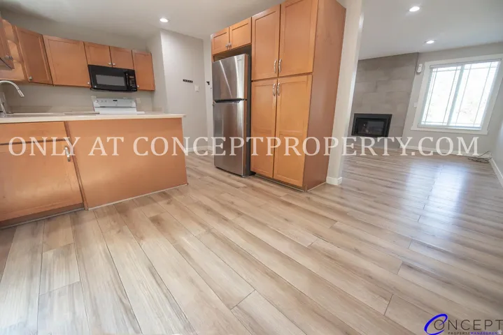 Wood-finish flooring throughout, kitchen with wood cabinetry and stainless steel refrigerator, white countertops, built-in microwave, and a fireplace with a light-colored tile surround