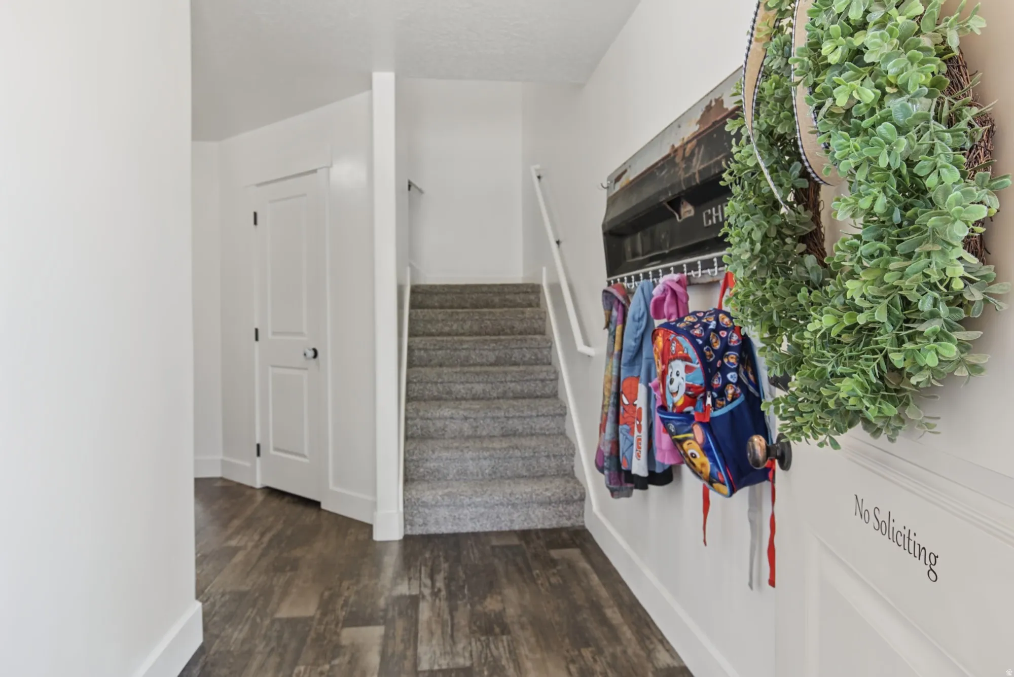 Wood-finish flooring, white paneled interior door with black hardware, and carpeted stairs with a white handrail