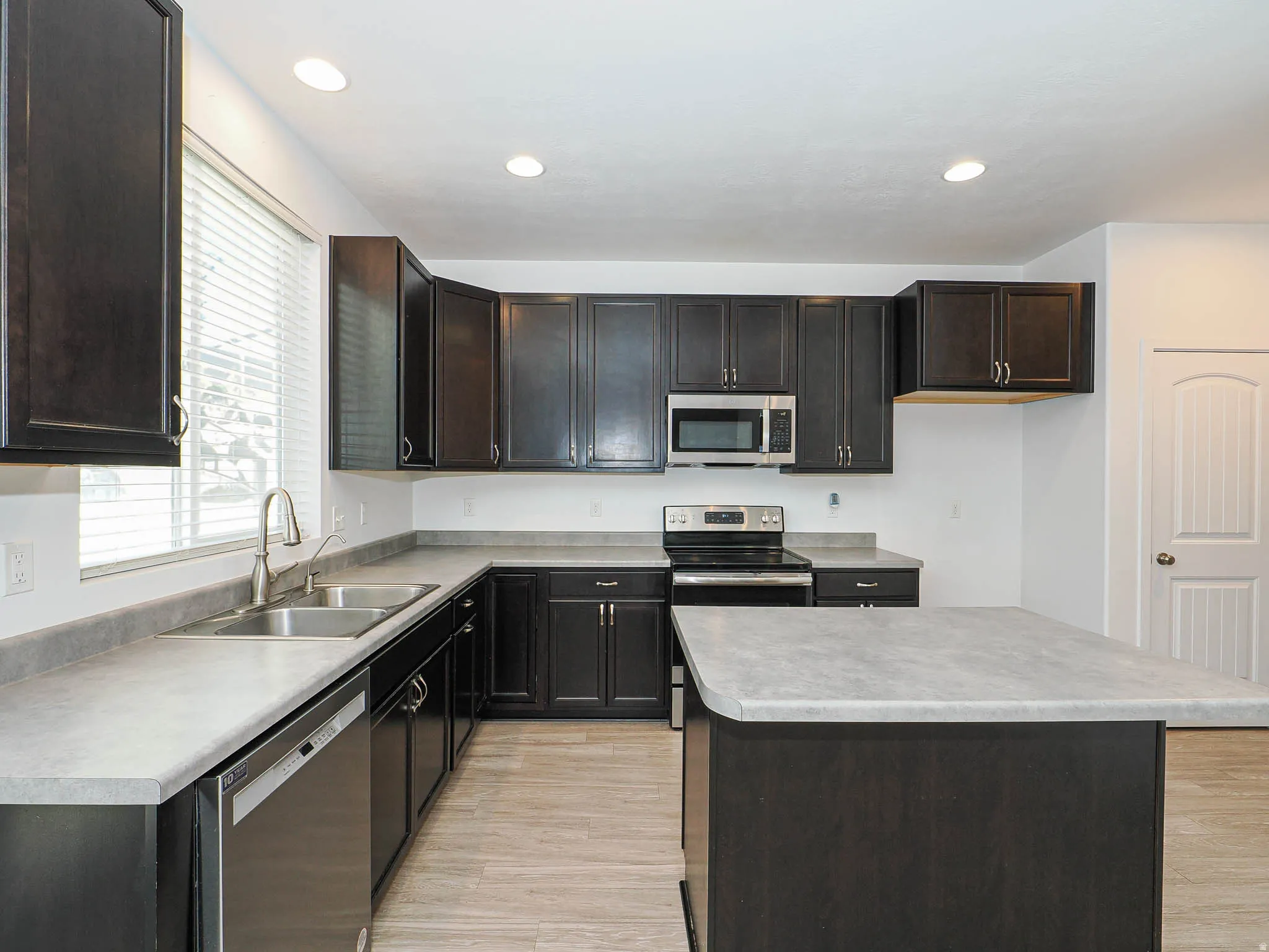 Expansive kitchen featuring dark wood cabinetry, light-toned countertops, stainless steel appliances, a dual basin sink, and an island with additional counter space