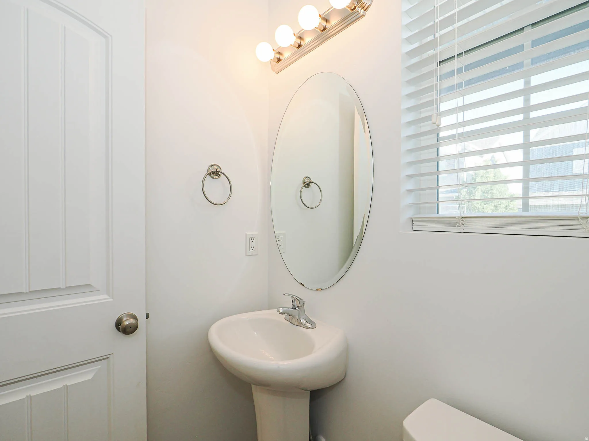 Pedestal sink with chrome faucet, oval mirror, and a three-bulb vanity light fixture