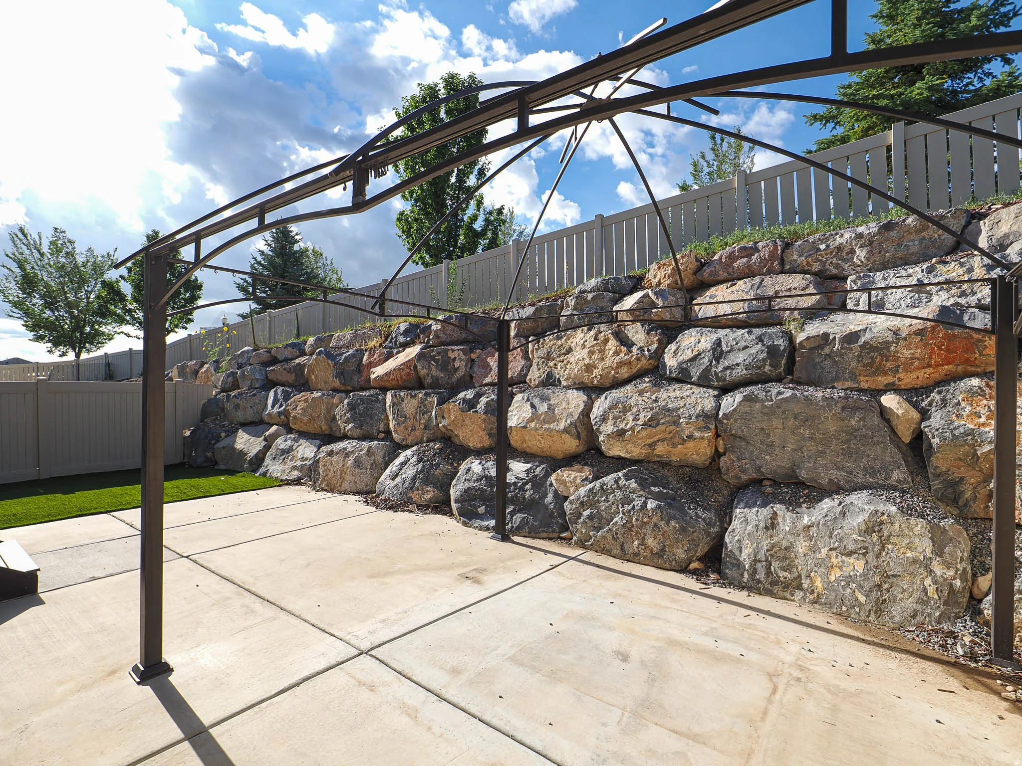 Expansive concrete patio area with a metal pergola frame, bordered by a substantial rock retaining wall and artificial turf