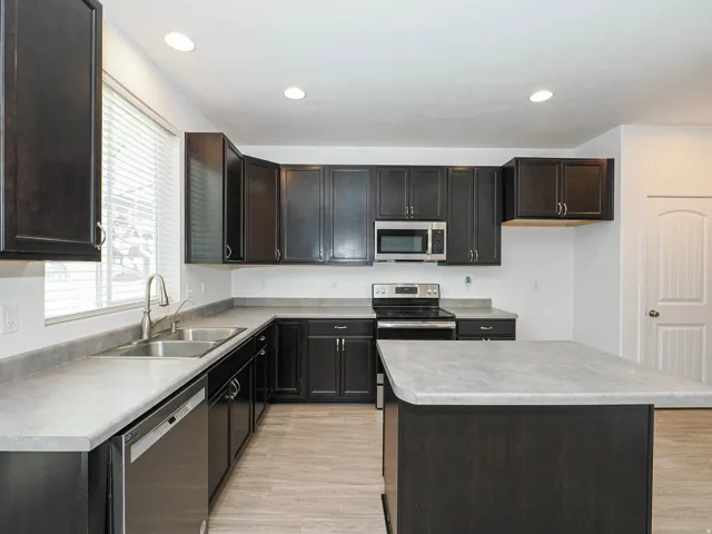 Expansive kitchen featuring dark wood cabinetry, light-toned countertops, stainless steel appliances, a dual basin sink, and an island with additional counter space
