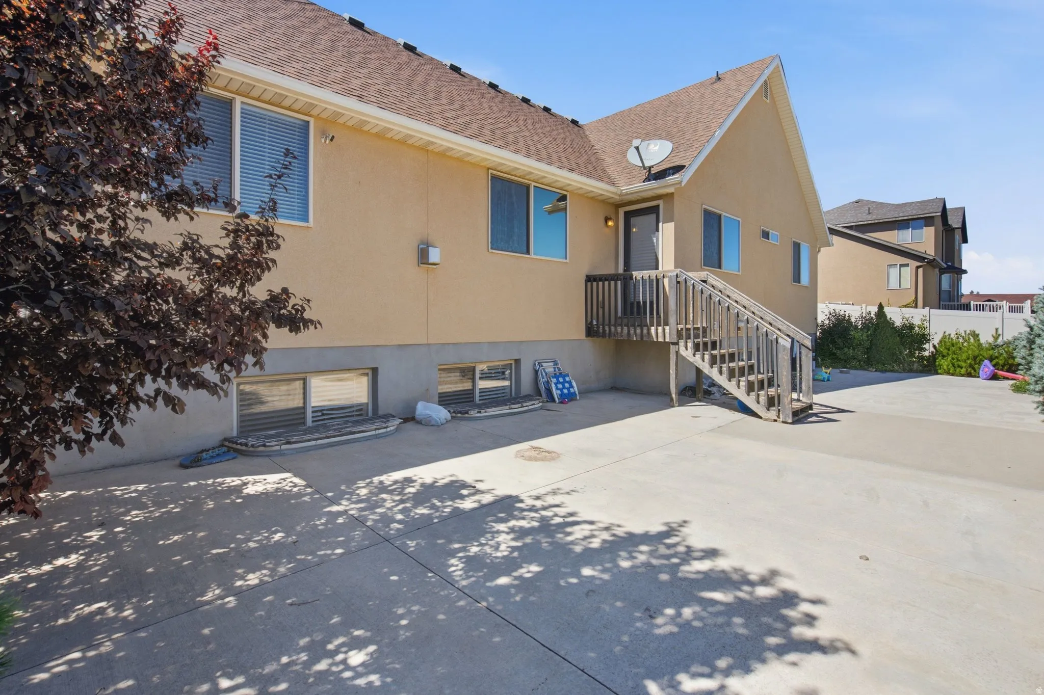 Exterior featuring a neutral stucco finish, multiple windows with blinds, a wood-finish deck with stairs, a concrete patio, and a shingle roof