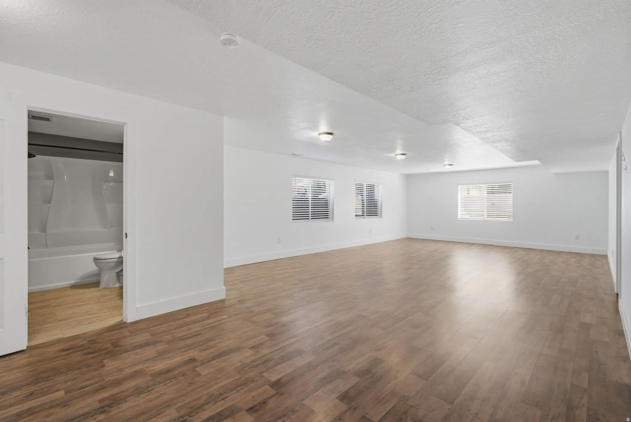 Expansive room with wood-finish flooring, white walls, multiple windows with blinds, and flush-mount ceiling lighting