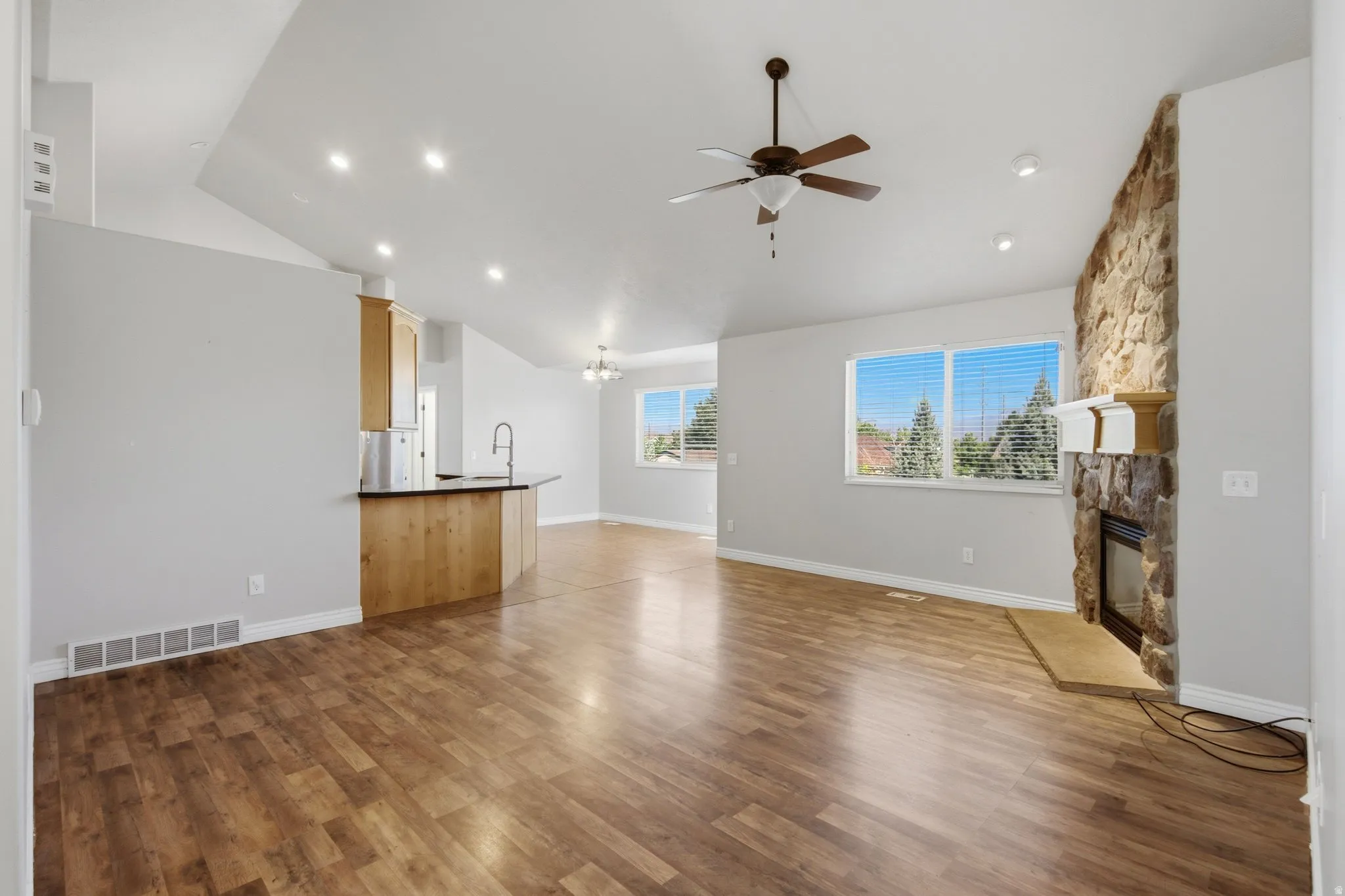 Vaulted ceilings, wood-finish flooring, stone fireplace with hearth, built-in kitchen peninsula, and ceiling fan with light fixture