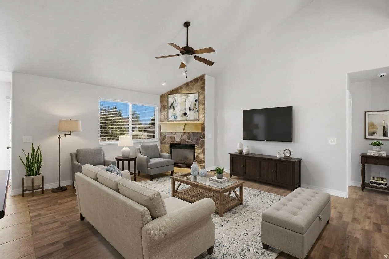 Vaulted ceiling with a ceiling fan, stone fireplace with a mantel, large windows, and wood-finish flooring