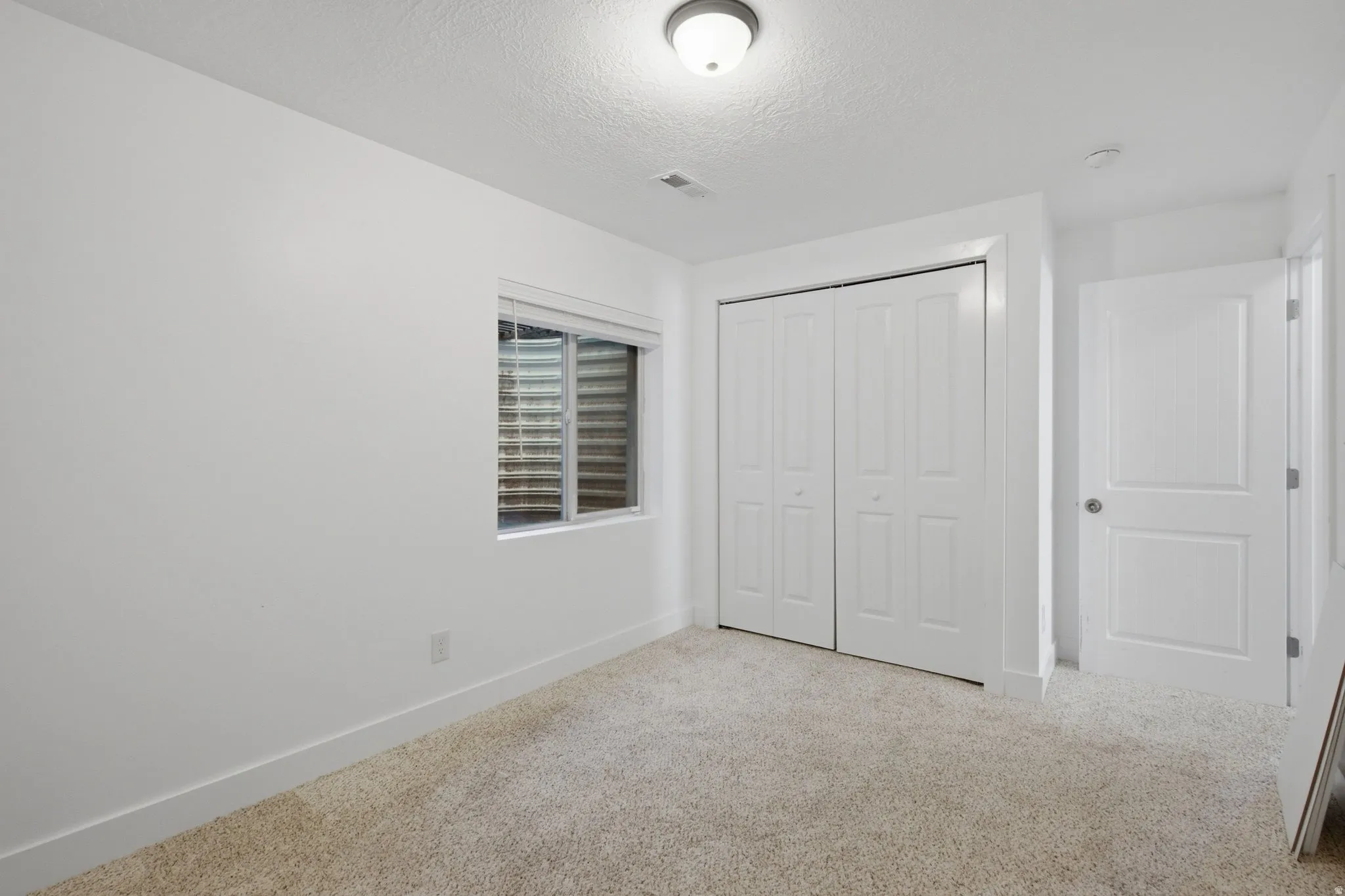 Recessed window with blinds, bi-fold closet doors, wall-mounted light fixture, white interior door, and carpeted flooring