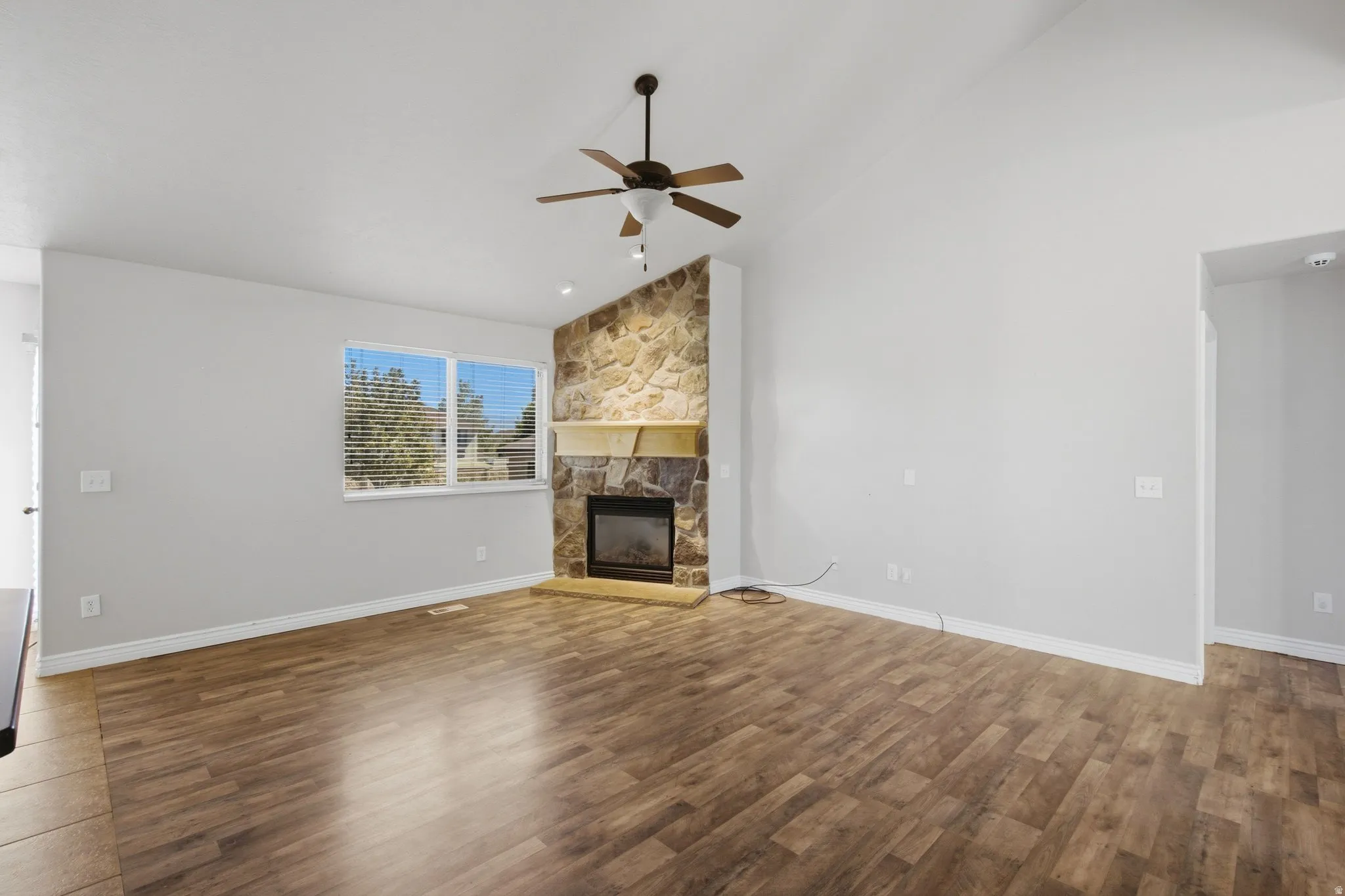 Vaulted ceiling with a ceiling fan, stone-clad fireplace with a wood mantle, wood-finish flooring, and a large window