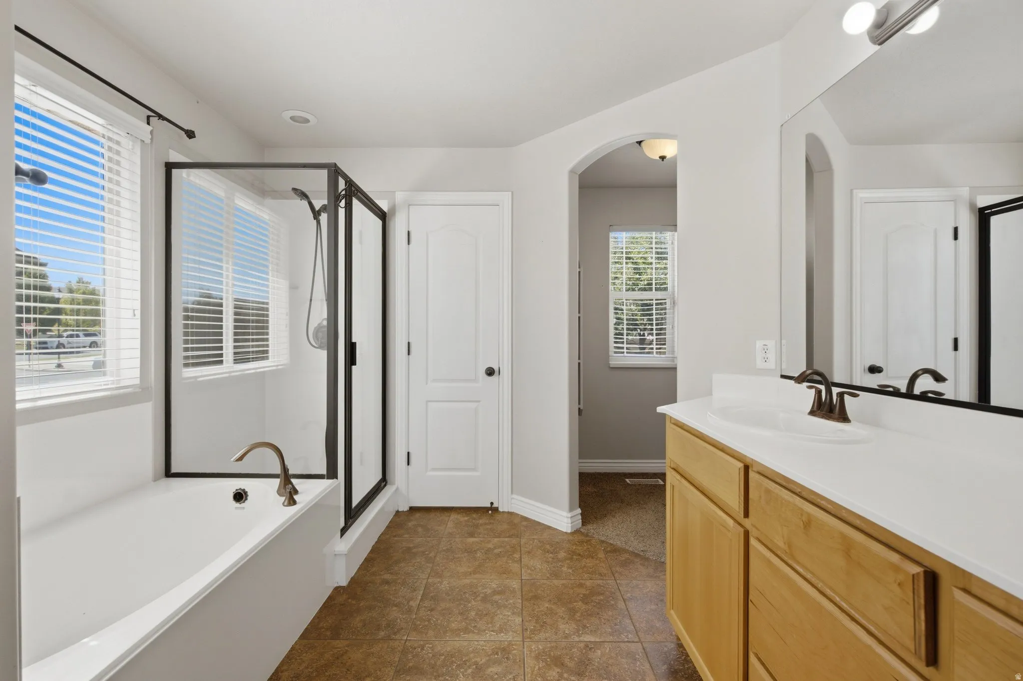 Expansive bathroom featuring a built-in tub, separate shower with a black-framed enclosure, dual windows with blinds, a vanity with a single sink, and tiled flooring