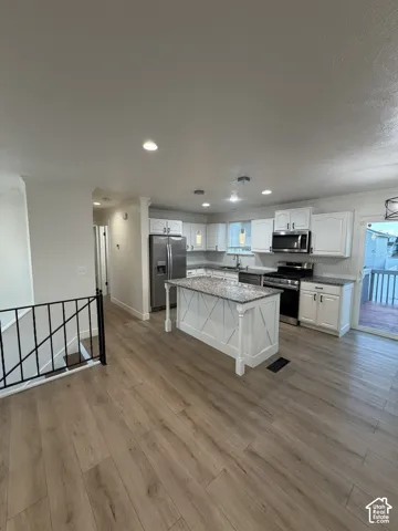 Kitchen featuring wood-finish flooring, white cabinetry, a center island with stone-finish countertops, stainless steel appliances, and black metal stair railing