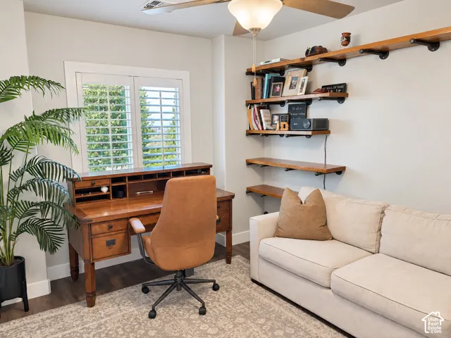 Window with plantation shutters, wood-finish flooring, built-in open shelving, and ceiling fan with light fixture