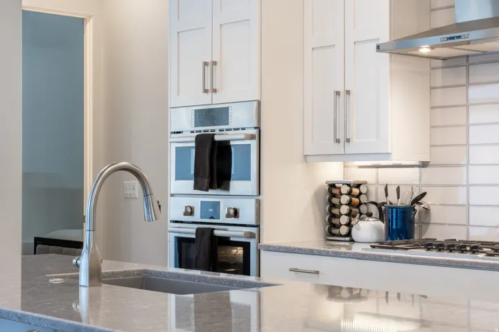 Stainless steel gooseneck faucet with an undermount sink, light gray countertop, white shaker cabinetry, and built-in double ovens