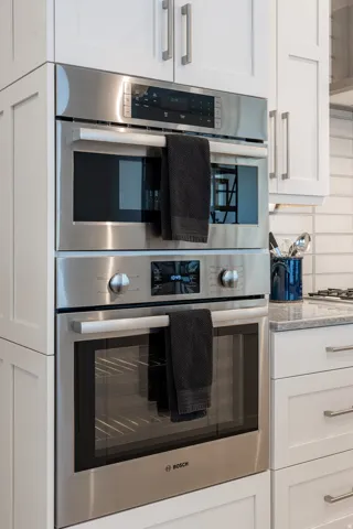 Built-in stainless steel appliances, stacked double ovens, white shaker cabinetry, and light-toned countertop