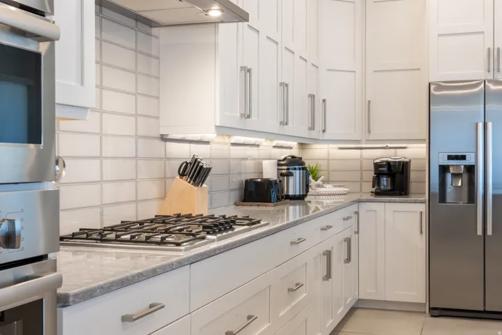 White shaker cabinetry with brushed nickel hardware, stainless steel appliances, stone-finish countertops, subway tile backsplash, and under-cabinet lighting