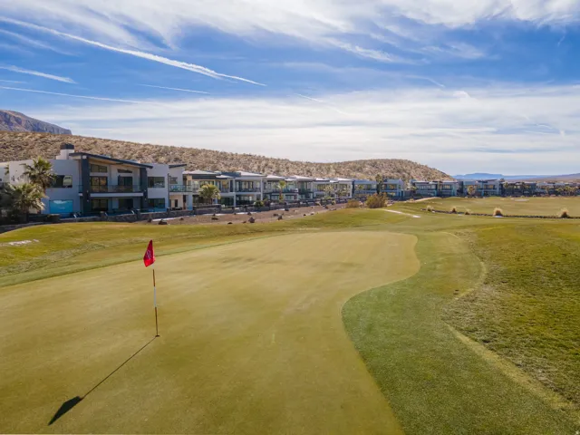 Manicured golf course green with flag, expansive golf course grounds, residential structures with multiple balconies, and distant natural desert landscape