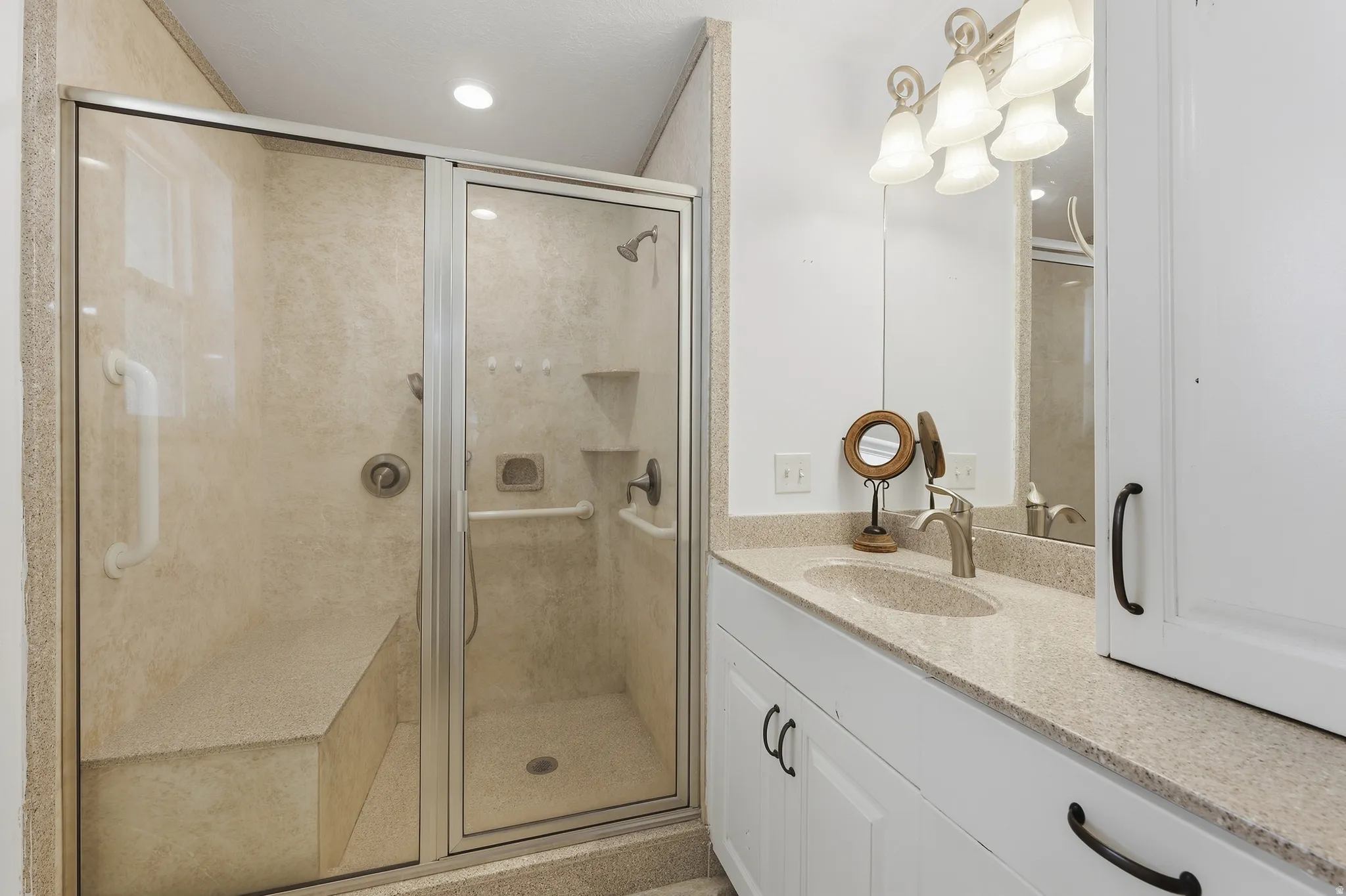 Walk-in shower with built-in bench and glass enclosure, recessed lighting, vanity with integrated sink, stone-finish countertop, and white cabinetry with dark hardware