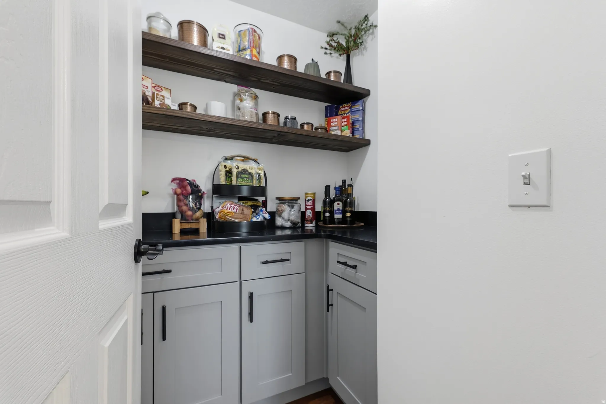 Pantry featuring open wood-finish shelving, dark countertops, and light gray cabinetry with dark hardware