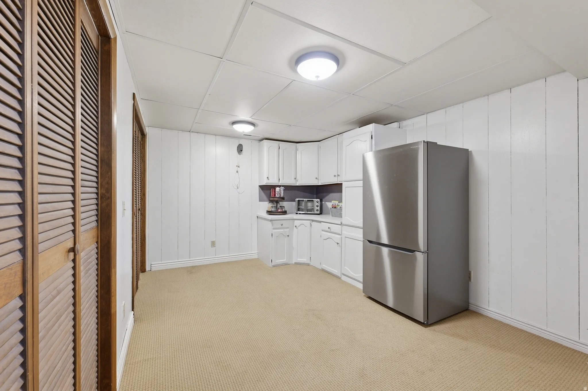 Recessed panel cabinetry, wood louvered bifold doors, stainless steel refrigerator, and drop ceiling with flush-mount lighting