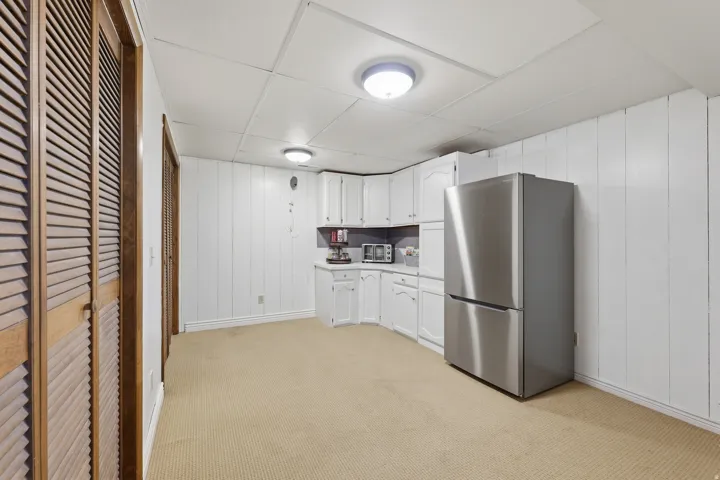Recessed panel cabinetry, wood louvered bifold doors, stainless steel refrigerator, and drop ceiling with flush-mount lighting