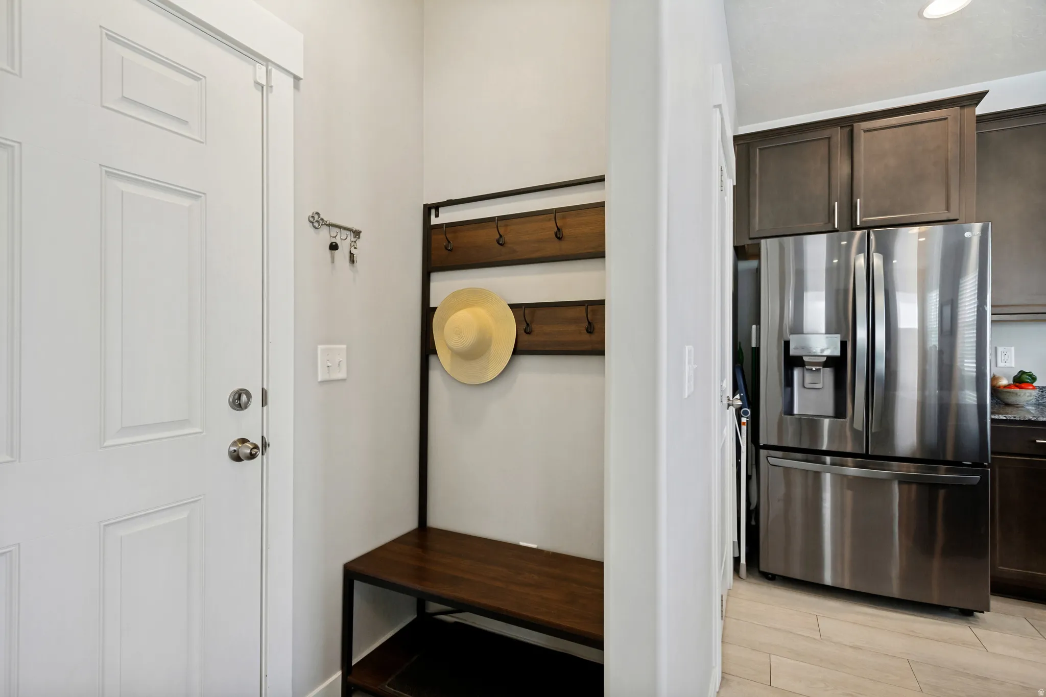 White paneled entry door, built-in coat rack with bench, stainless steel French door refrigerator, dark wood-finish cabinetry, and light wood-finish flooring
