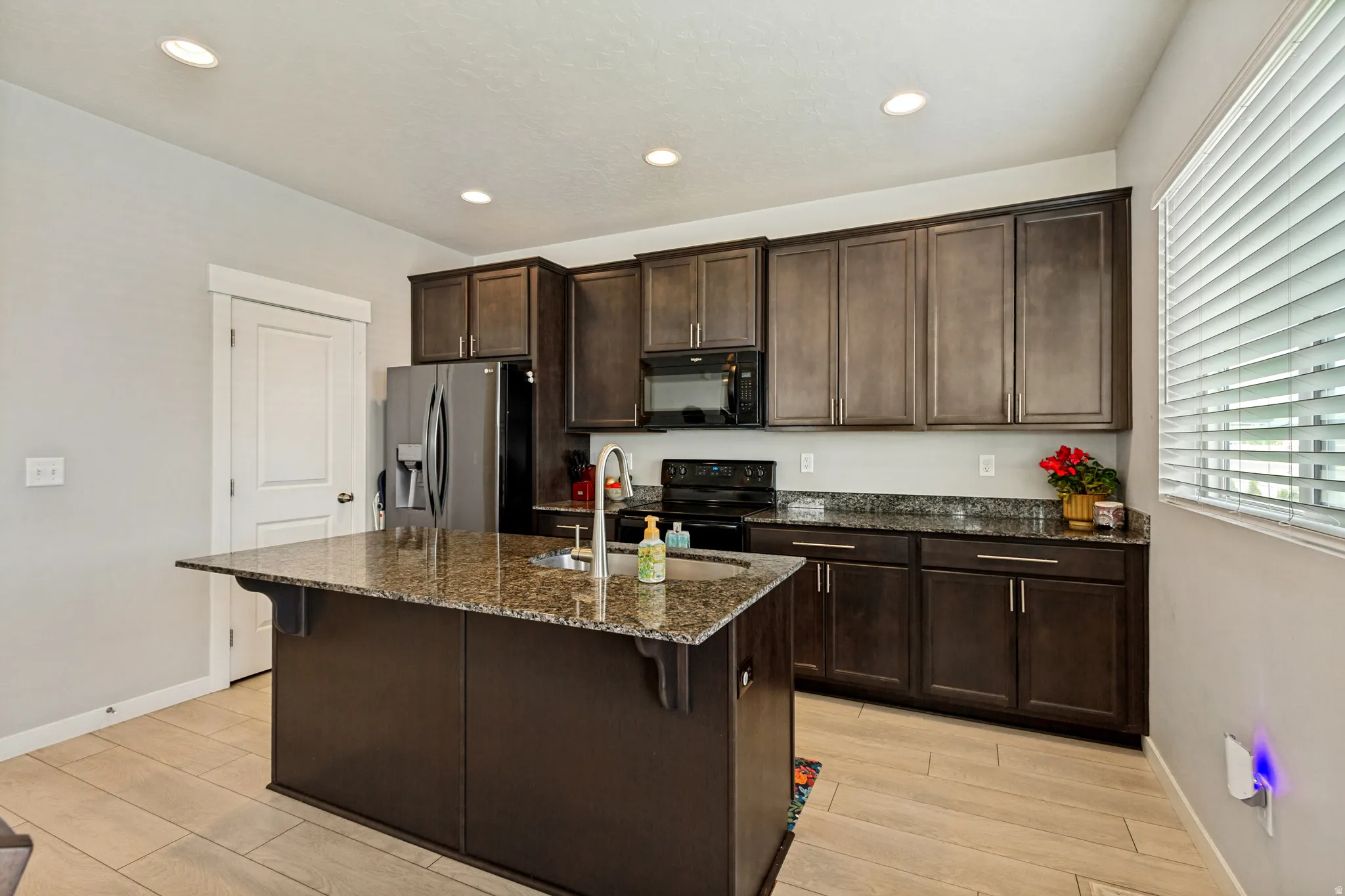 Kitchen featuring a central island with a sink, stainless steel appliances, dark wood-finish cabinetry, and granite countertops