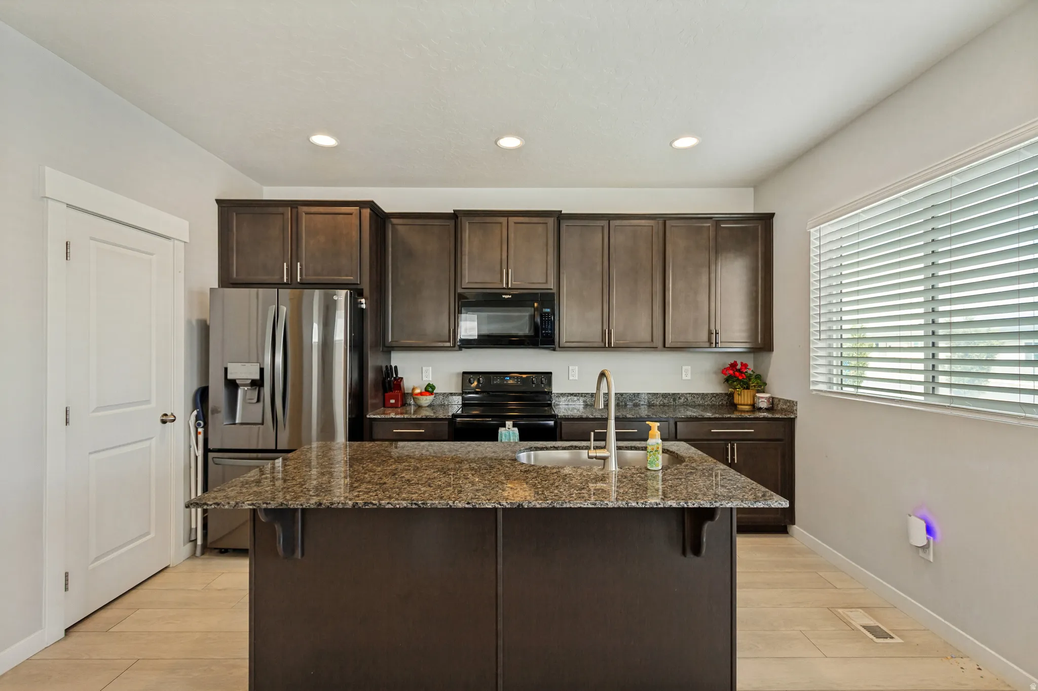 Kitchen featuring a large island with sink, dark wood cabinetry, stainless steel refrigerator, black range, and recessed lighting