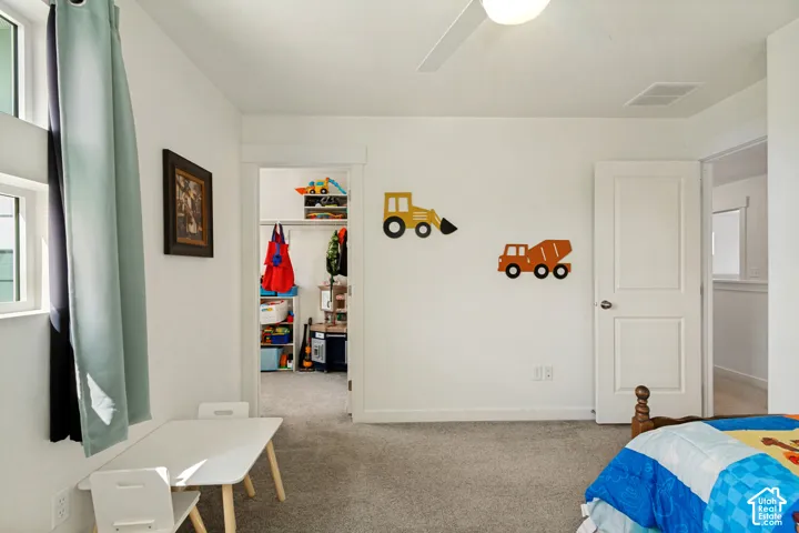 White painted walls with gray carpet flooring, an open doorway leading to a walk-in closet, and a ceiling fan with light fixture