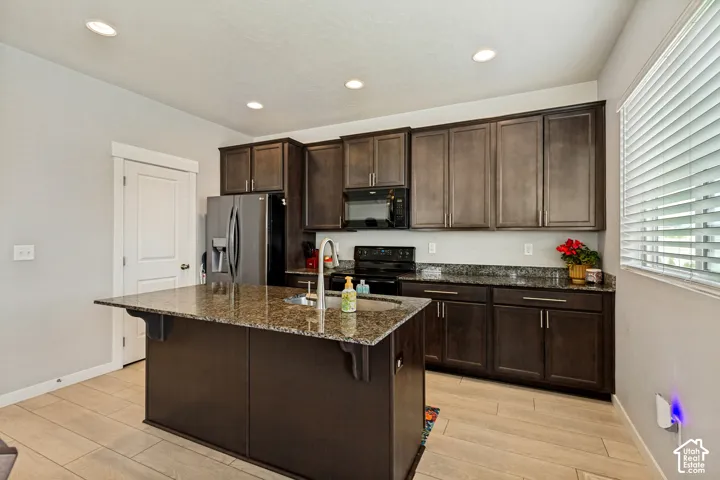 Kitchen featuring a central island with a sink, stainless steel appliances, dark wood-finish cabinetry, and granite countertops